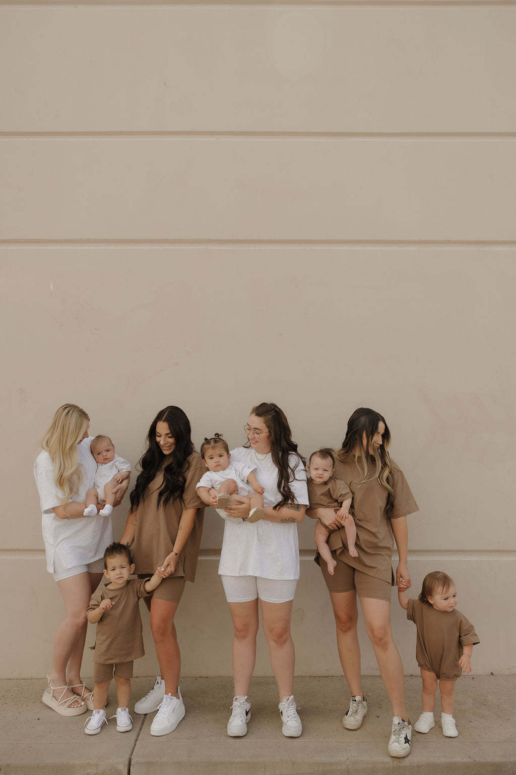 Four women and five small children pose together against a beige wall. The kids wear forever french baby’s stylish, comfortable Child Short Set | Patina, all looking relaxed and happy in coordinated white and brown outfits.