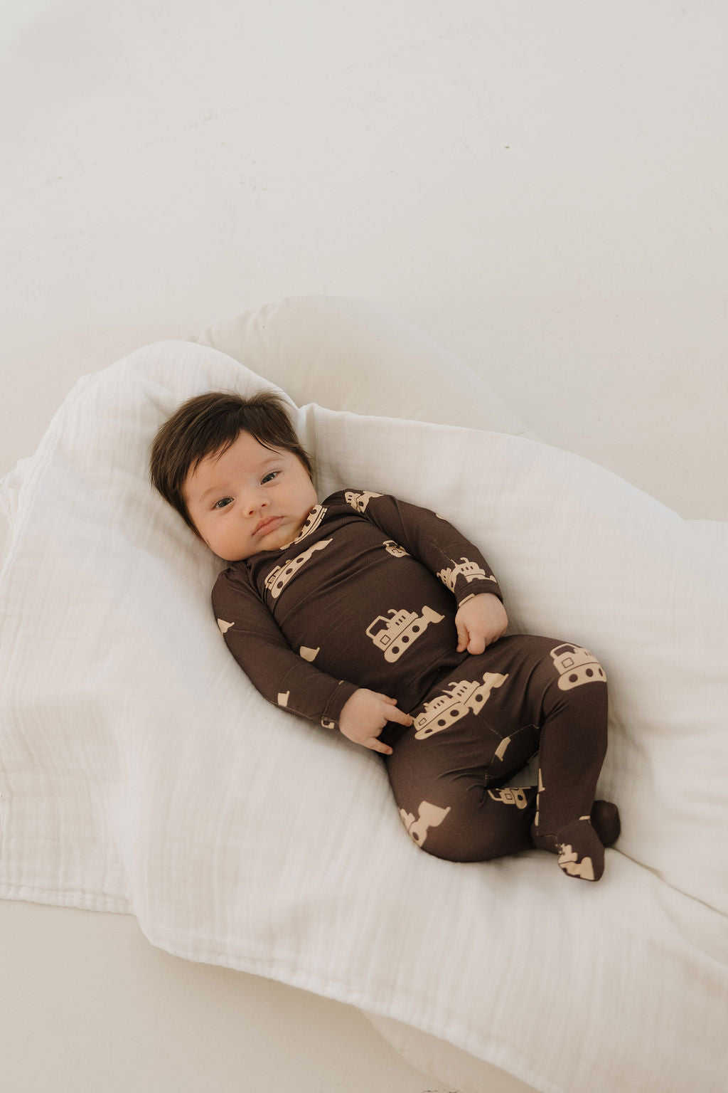 A baby in the forever french baby Sleepy Time Set | Bulldozer— a breathable brown onesie with light vehicle patterns— lies calmly on a white blanket, gazing upward.