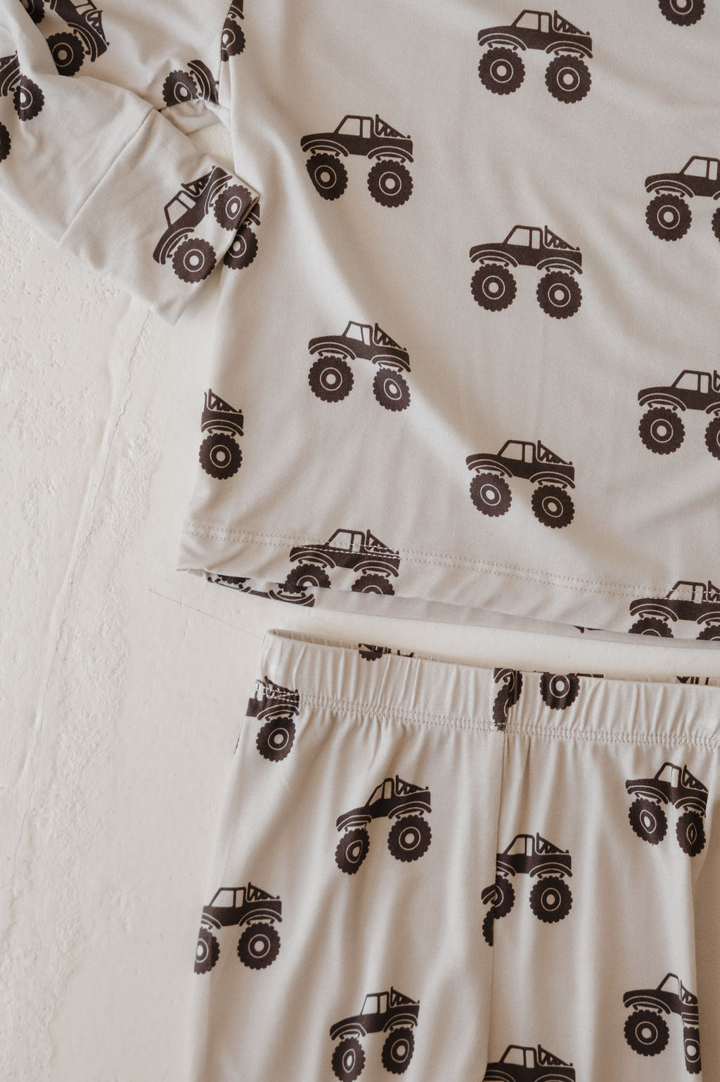 A close-up of the forever french baby Bamboo Footless Sleepy Time Set | Monster Truck, a hypo-allergenic, cream pajama set with black monster truck prints on the long-sleeve top and pants, laid out on a textured surface.