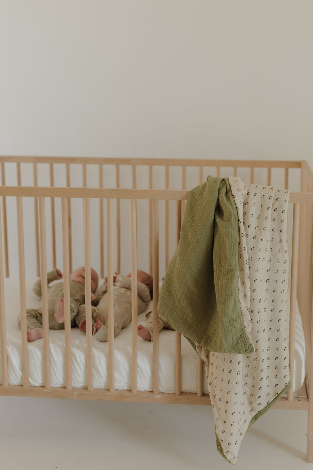 A wooden crib with a white mattress holds two babies, one in beige and the other in green. A forever french baby Muslin Quilt | Flora and a patterned blanket drape over the rail, adding charm to the nursery against a plain light wall.