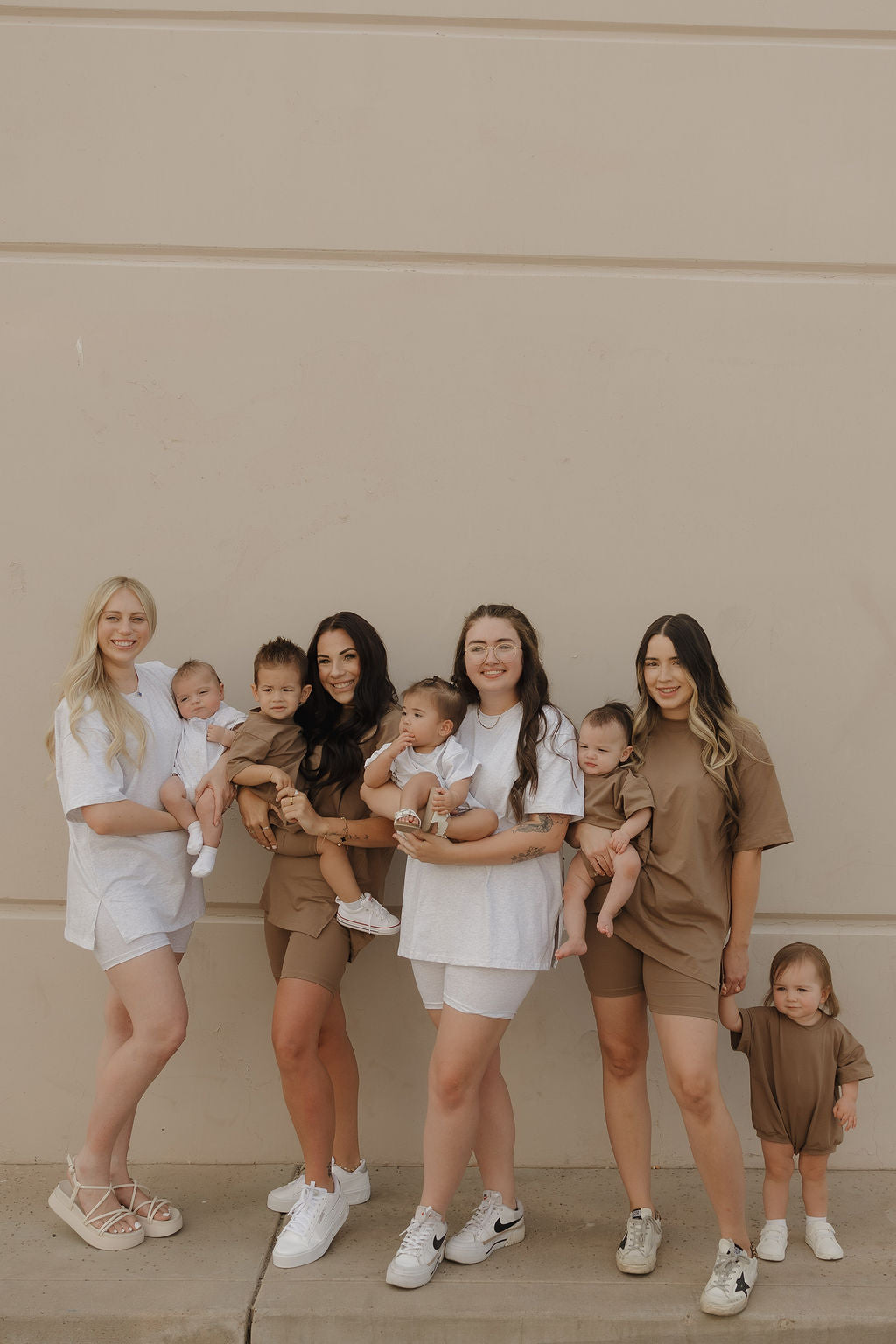 Four women and their children gather by a beige wall, wearing matching forever french baby Short Sleeve Romper | Patina in neutral tones with handy snap closures. One toddler stands next to the group, holding a woman's hand.