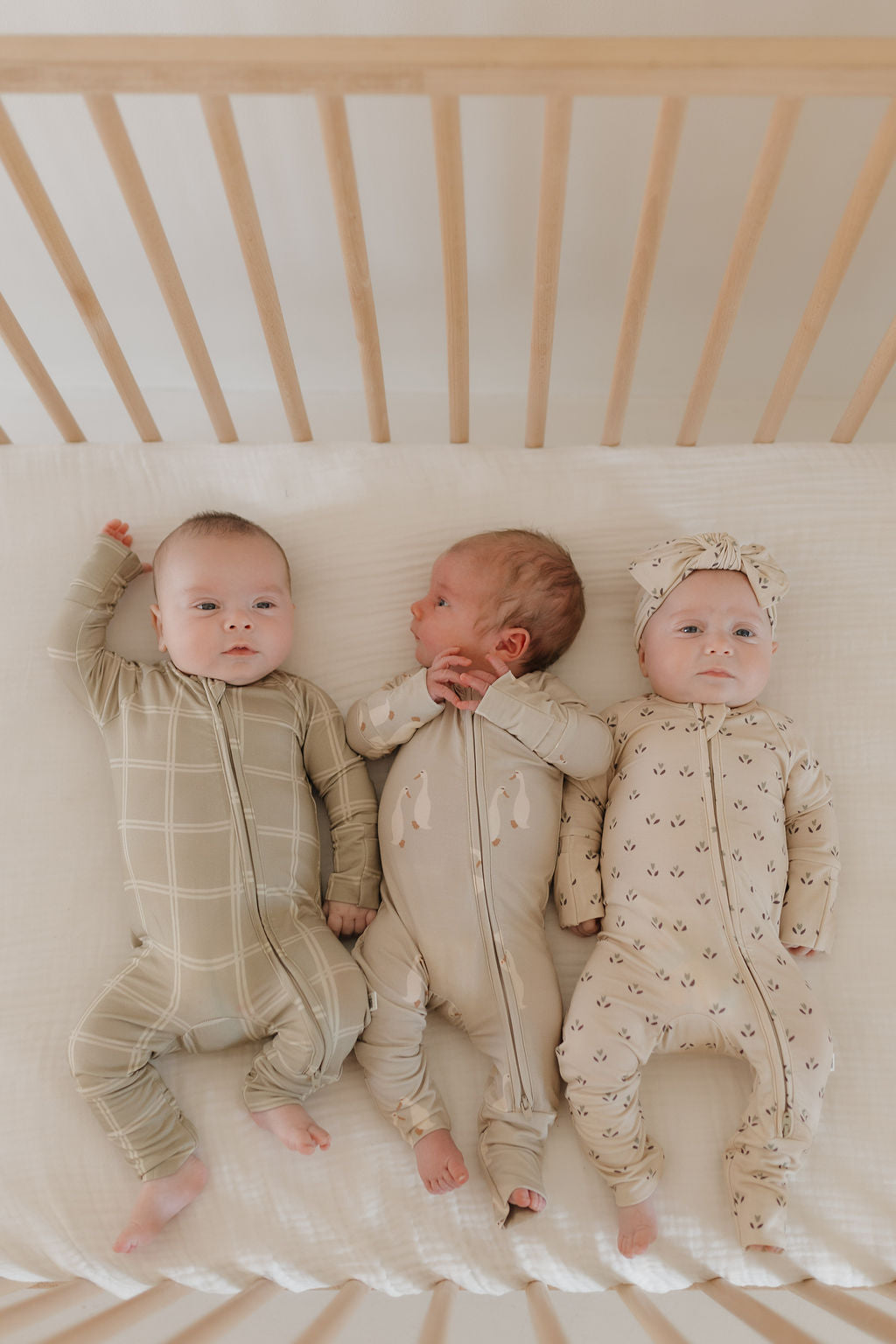 Three babies rest side by side in a crib with wooden bars. The one on the left wears Bamboo Zip Pajamas in Linen Grid by forever french baby, the middle sports animal prints, and the right one is dressed in a patterned onesie with a matching headband.