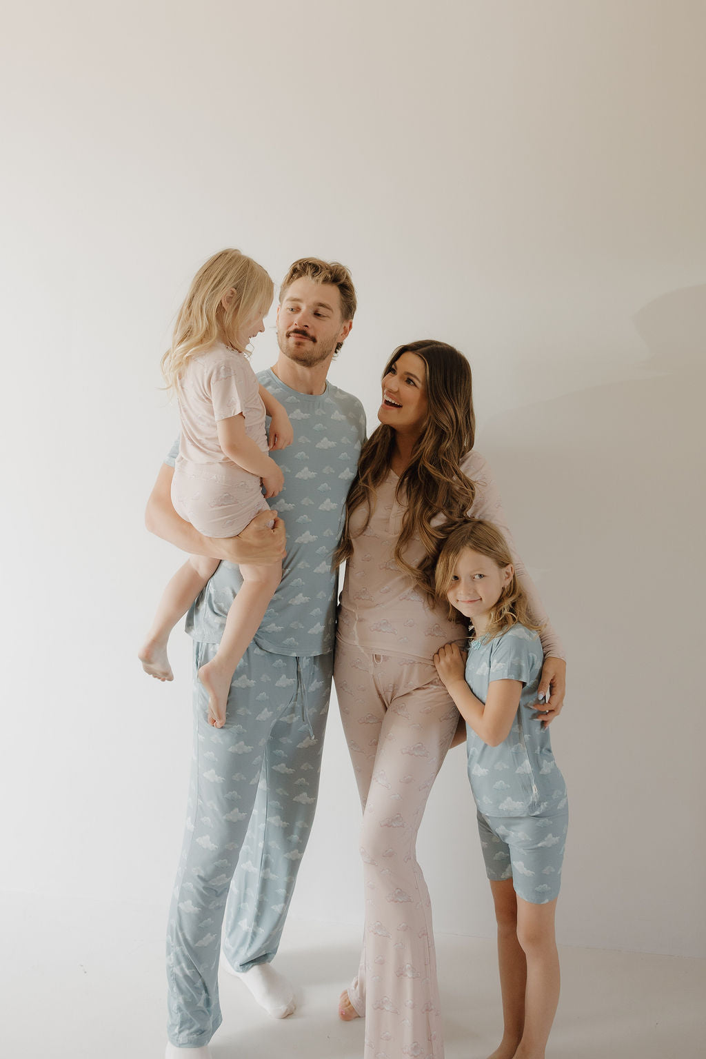 A smiling family of four wears matching Kendy 3 Bamboo Short Set | Duke pajamas. Barefoot, the father holds a young girl while the mother hugs their older daughter, all posing together against a plain background.