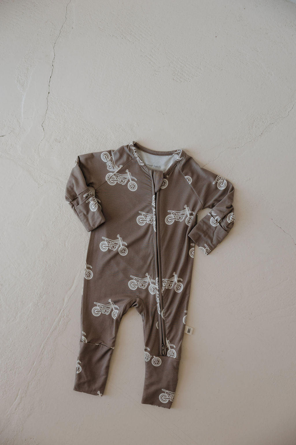 The Bamboo Zip Pajamas | Dirt Bike by forever french baby is a brown, hypoallergenic baby onesie with long sleeves, a front zipper, and a white bicycle pattern, shown laid flat on a light textured surface.