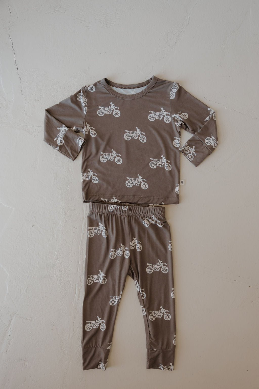 The forever french baby Bamboo Footless Sleepy Time Set | Dirt Bike is a two-piece brown children’s pajama set with long sleeves and pants, featuring a white dirt bike print. Made from hypo-allergenic bamboo for cozy comfort and laid flat on a textured surface.