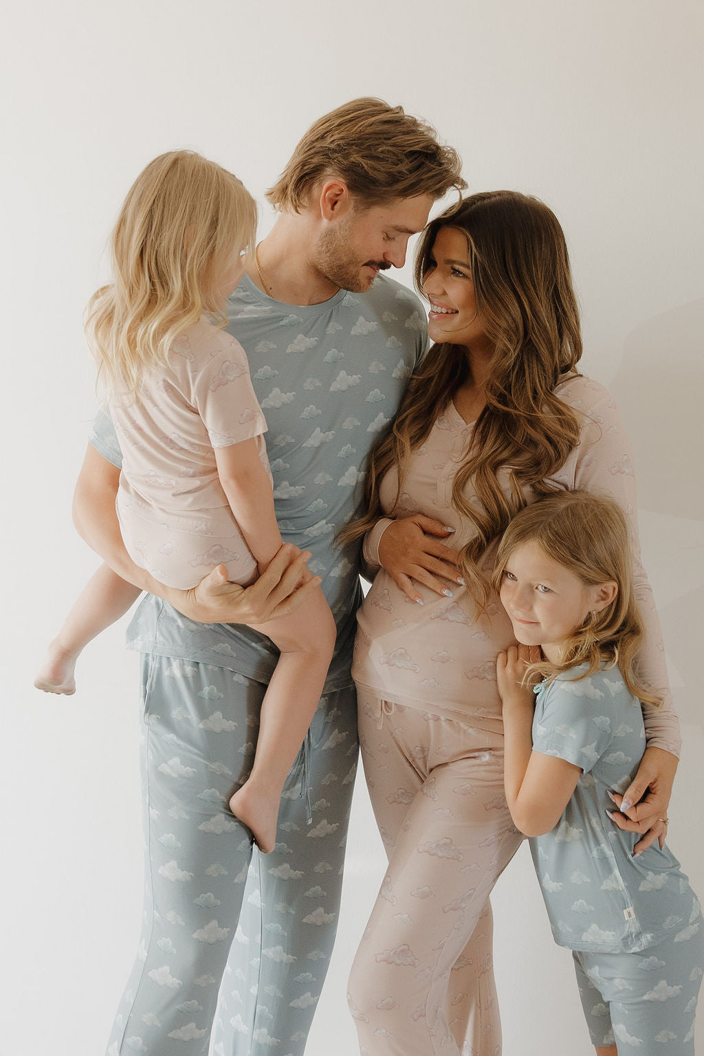 A smiling family of four wears matching Kendy 3 "Bamboo Short Set | Ruby" pajamas, featuring hypo-allergenic comfort and stylish cloud patterns as the father holds one child and the mother hugs the other.