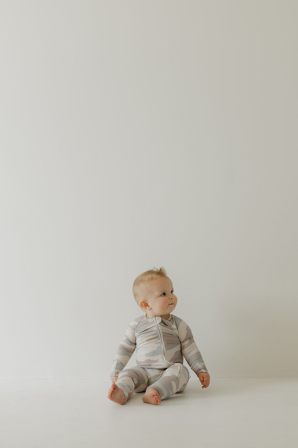 In a minimalistic setting, a baby sits peacefully on the floor, wearing comfortable Bamboo Zip Pajamas in Camo by forever french baby. The child's expression is calm, with hands resting gently on the floor and bare feet relaxed.