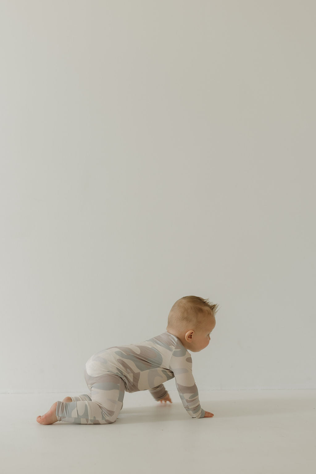 A baby wearing breathable Bamboo Zip Pajamas in Camo by forever french baby crawls on a smooth, light floor against a plain wall, facing away from the camera. The scene is minimalistic and softly lit.