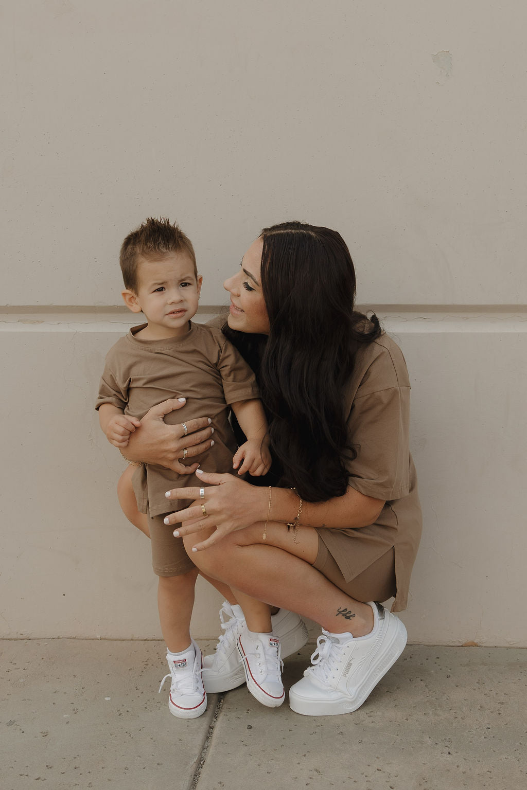 A woman with long dark hair crouches next to a young boy, both in matching forever french baby Child Short Set | Patina—biker shorts and oversized tees paired with white sneakers. She smiles at him, holding him gently against a plain light-colored wall.