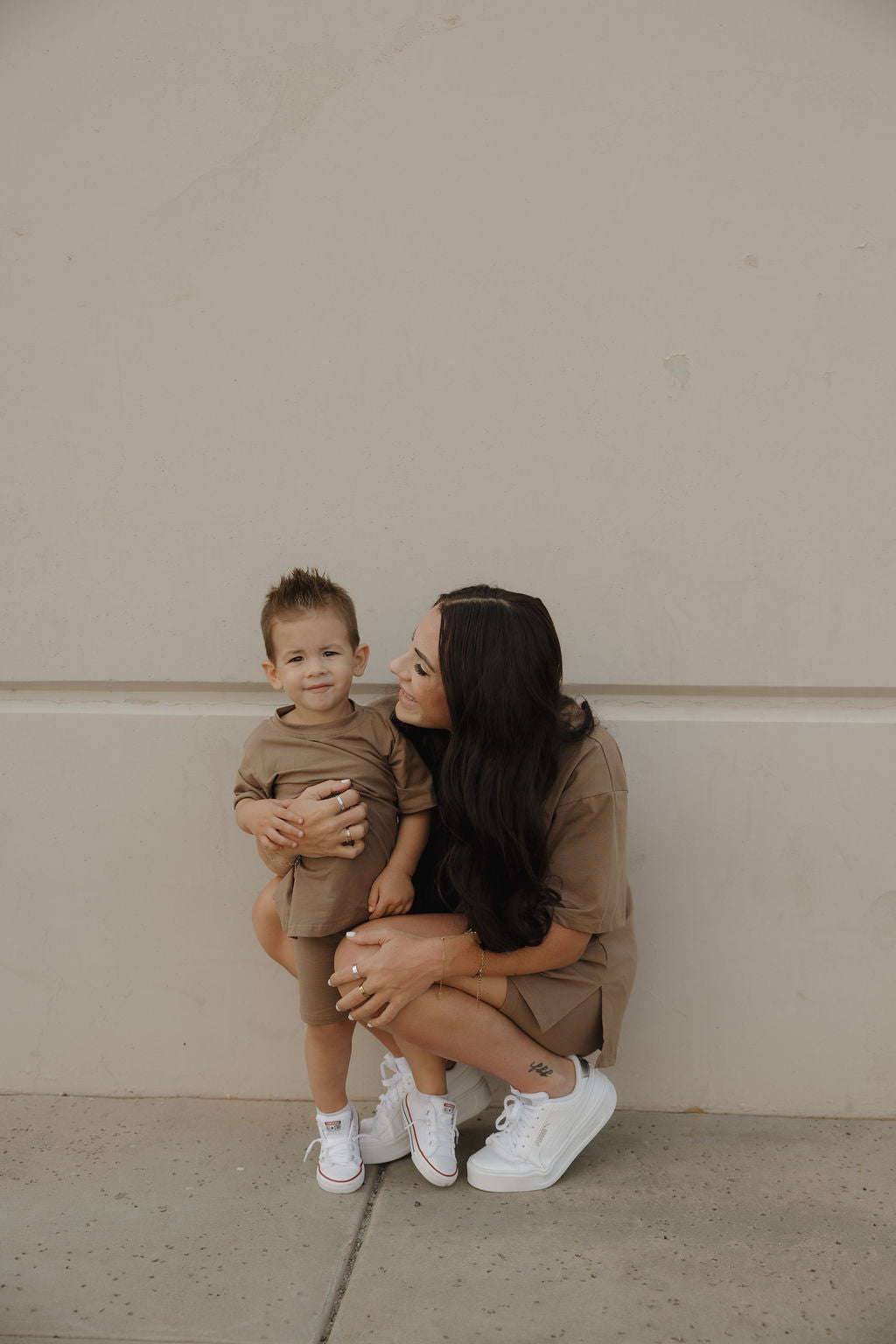 A woman with long dark hair crouches next to a young boy, both in matching forever french baby Women's Short Set | Patina and white sneakers, sharing a smile as they pose against a plain beige wall.