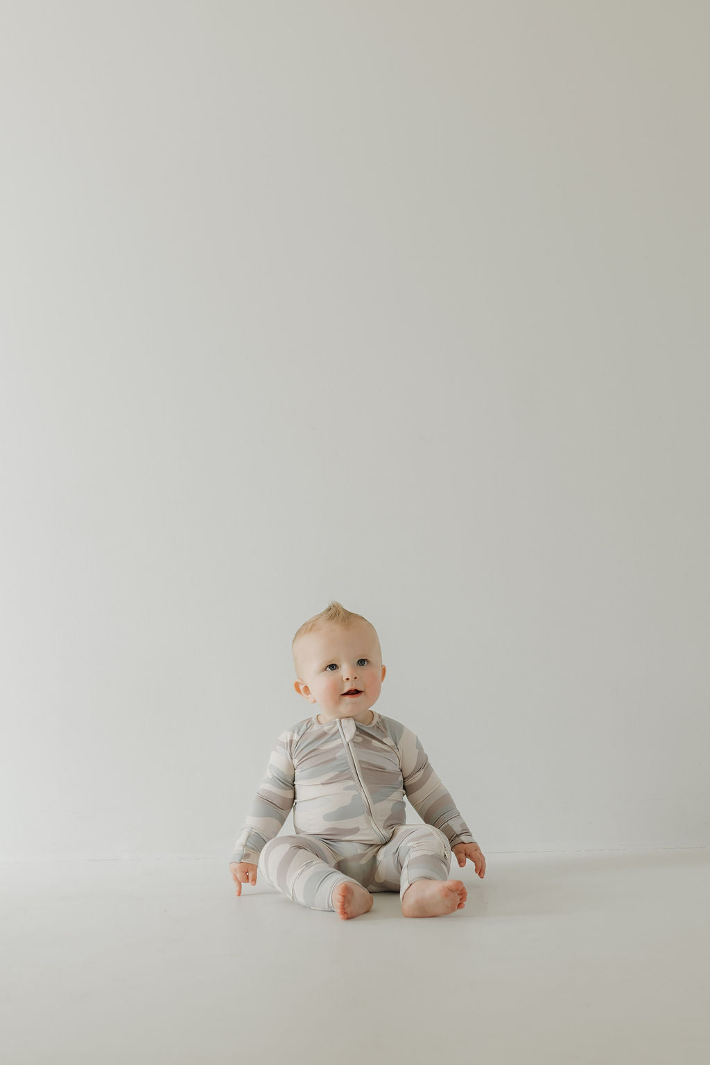 A baby in Bamboo Zip Pajamas | Camo by forever french baby sits on a plain white floor against a minimalist, pale background. With a curious expression, the baby faces forward, comfortably dressed in hypo-allergenic clothing perfect for sensitive skin.