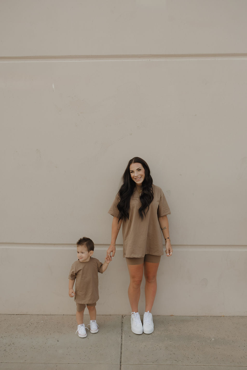 A woman and young child stand hand in hand, smiling in matching forever french baby short sets (Patina). The woman’s oversized tee and biker shorts create a modern, casual vibe against the beige wall. Both wear white sneakers.