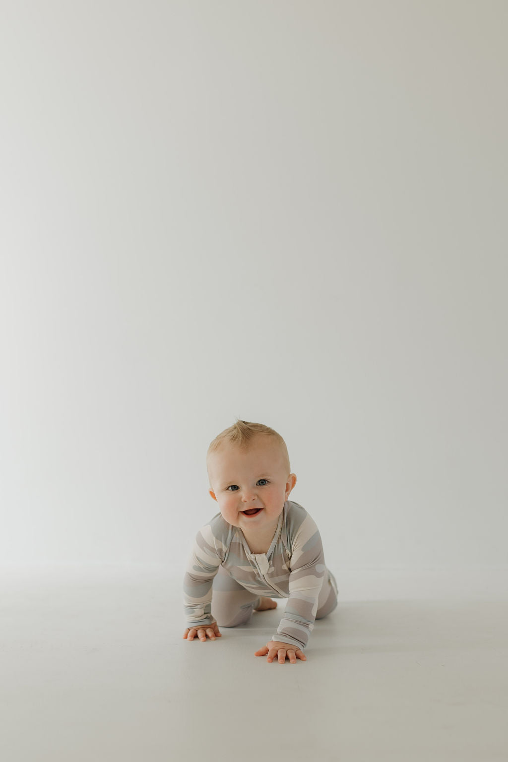 A baby in bamboo camo pajamas is crawling on a white floor against a plain white background, enjoying the comfort of breathable sleepwear by forever french baby.