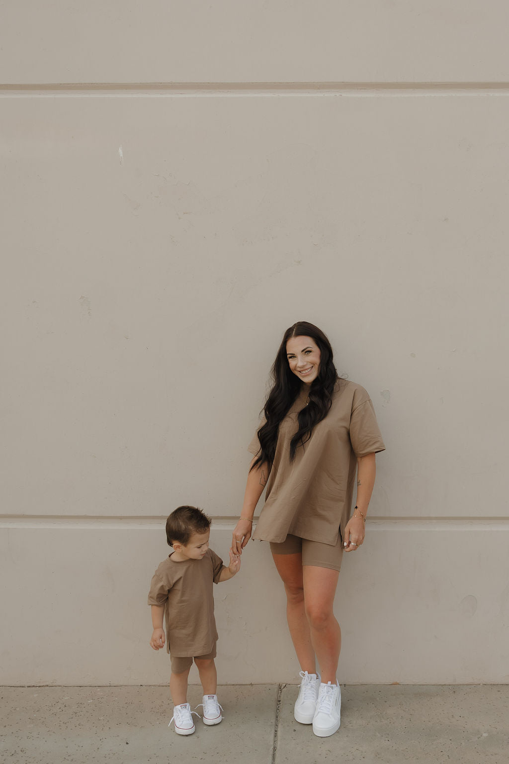 A smiling woman and a young child stand against a beige wall, both wearing matching forever french baby Women's Short Set | Patina and white sneakers, while the woman holds the child's hand.