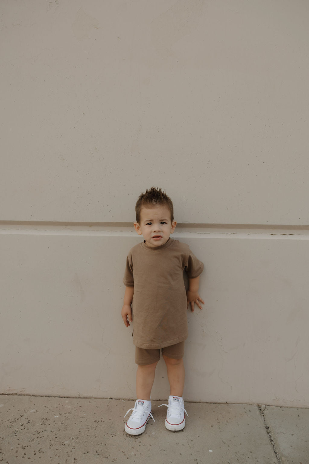 A young child with short brown hair wears the forever french baby Child Short Set in Patina, paired with white sneakers and stands against a plain beige wall, looking at the camera with a neutral expression.