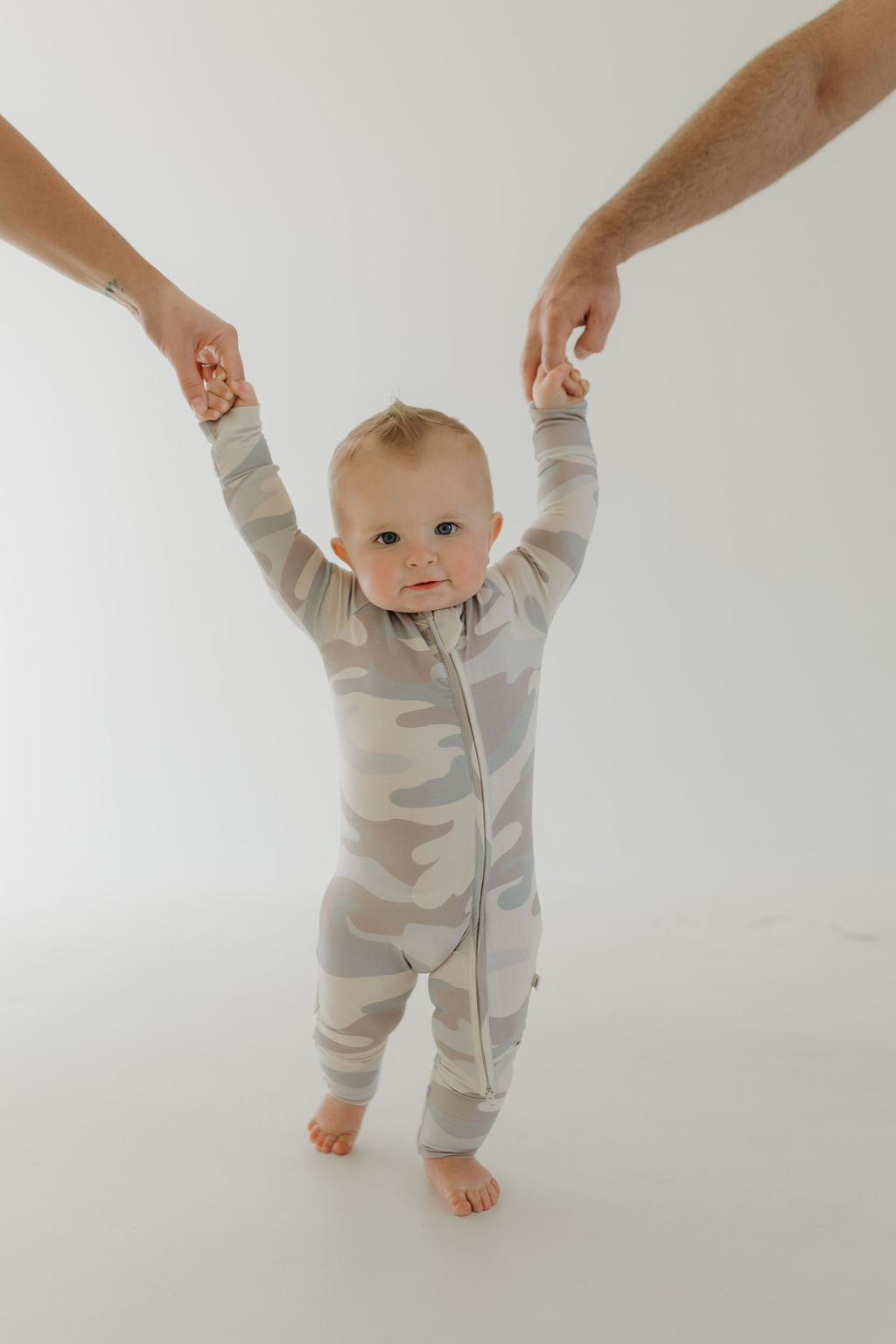 In a plain white setting, a baby in forever french baby's Bamboo Zip Pajamas | Camo, crafted from hypoallergenic fabric, holds hands with two adults. With a small smile, the child looks forward, radiating comfort. Only the adults' arms are visible.