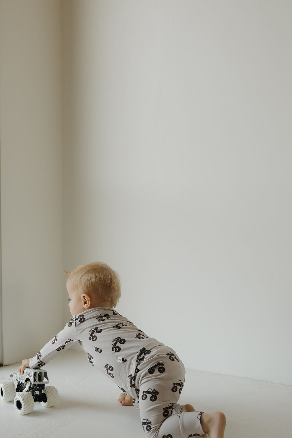 A young child in the forever french baby Bamboo Footless Sleepy Time Set | Monster Truck crawls on the floor, reaching for a toy truck near a plain white wall.