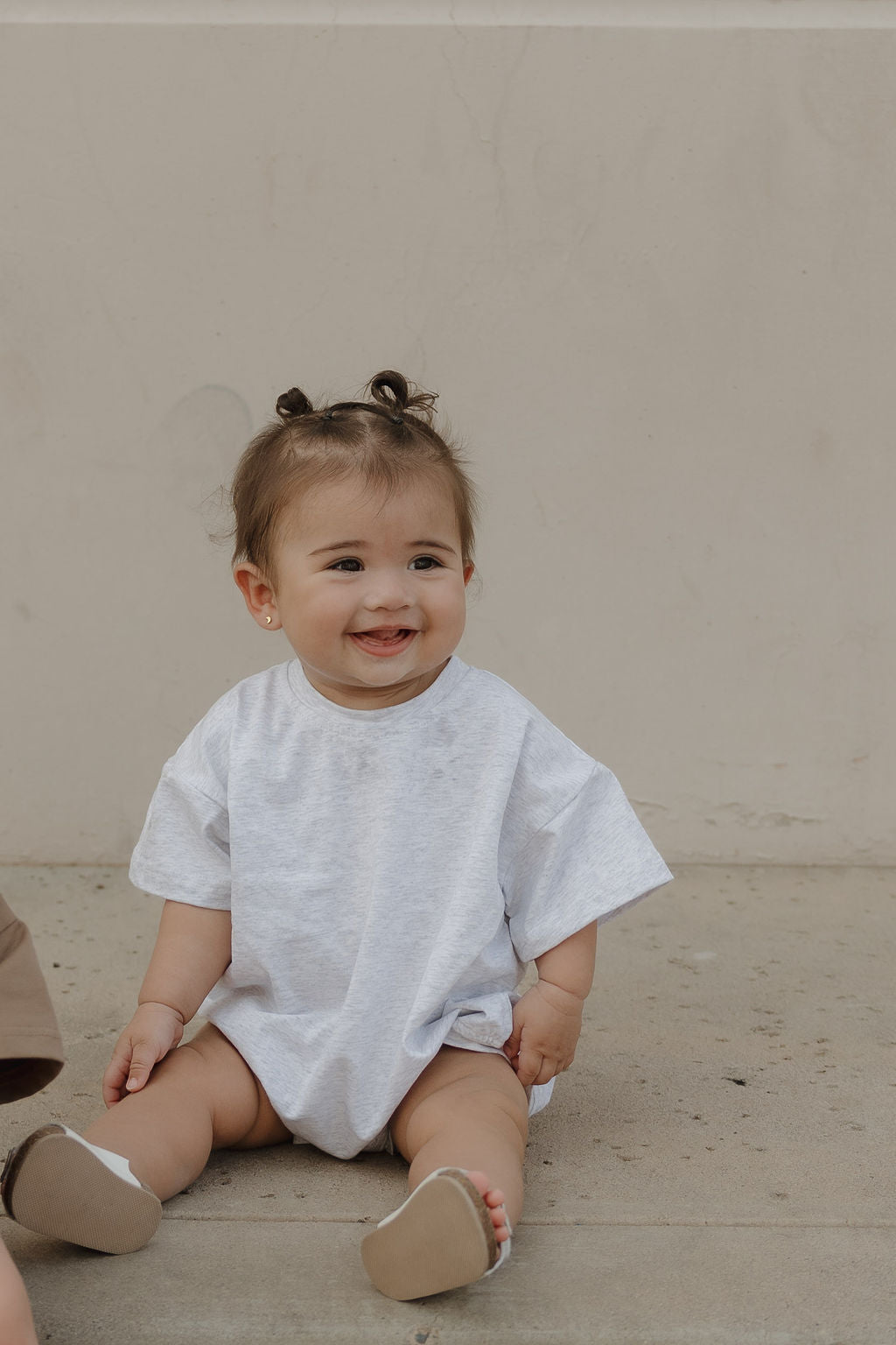 A smiling baby with short brown hair in pigtails sits on concrete, wearing the forever french baby Short Sleeve Romper | Heather and light shoes, against a plain light beige wall.