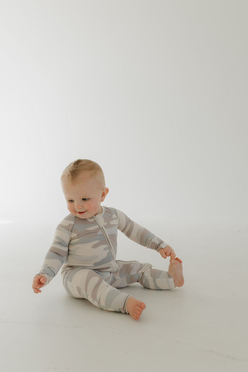 A baby, dressed in Forever French Baby's gray camo-patterned Bamboo Zip Pajamas, sits on a light-colored floor with a slight smile. The hypo-allergenic fabric complements the minimalistic white background, enhancing the baby's playful expression.
