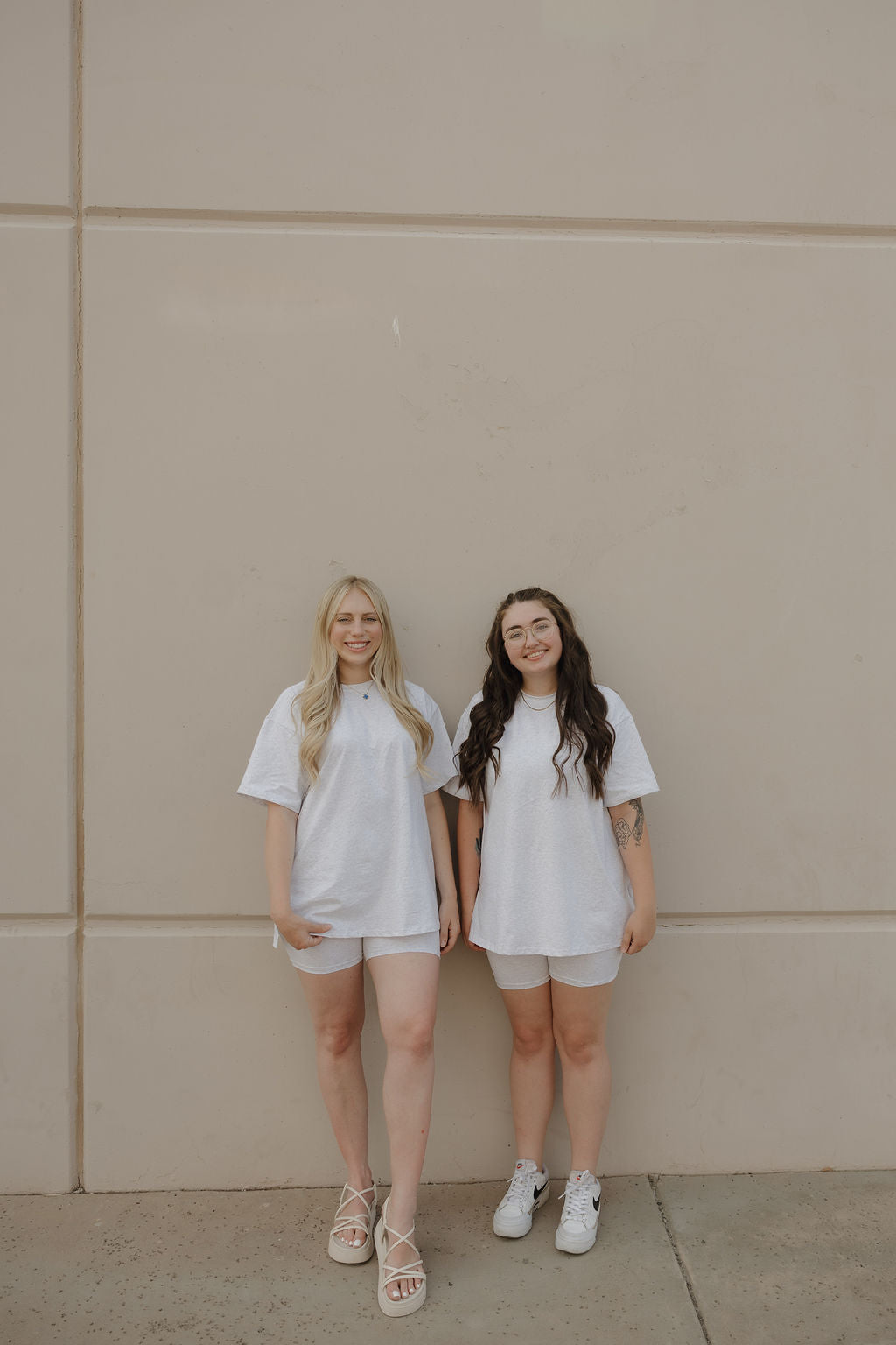 Two women smile in front of a beige wall, both wearing forever french baby Women's Short Set | Heather. One pairs hers with sandals and long blonde hair; the other wears sneakers and has long dark hair.