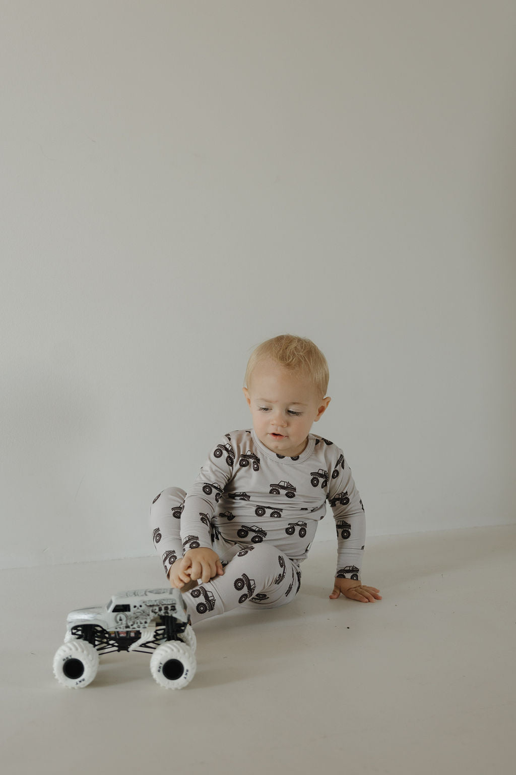 A young child wearing the Bamboo Footless Sleepy Time Set | Monster Truck by forever french baby sits on a plain light background, reaching forward to play with a toy monster truck.