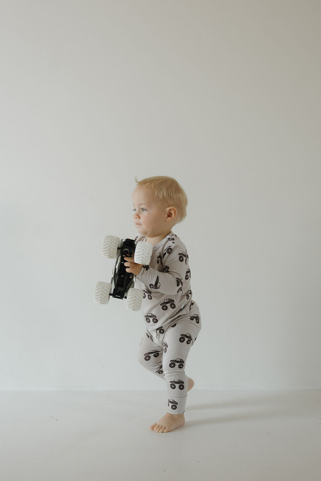 A toddler in the forever french baby Bamboo Footless Sleepy Time Set | Monster Truck stands barefoot on a white floor, holding a large toy truck. The monster truck print adds playful charm to the scene against a plain white background.