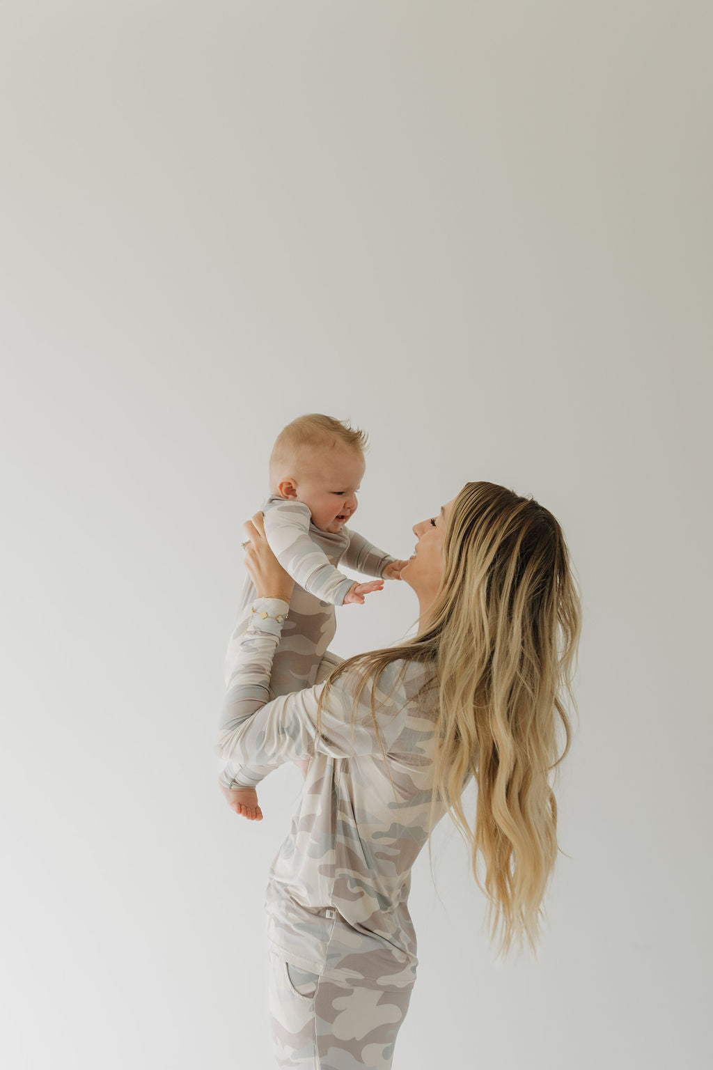 A smiling baby and a woman with long blonde hair joyfully wear matching forever french baby Women's Bamboo Pajama in Camo, highlighting the hypo-allergenic comfort. The plain light background enhances the image's soft, minimalist feel.