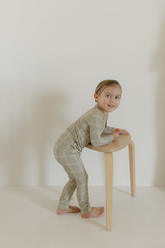 A playful child wears forever french baby's Bamboo Two Piece Pajamas in Linen Grid, standing on white flooring and leaning on a wooden stool. With a red wristband and a smile, the child poses against a white minimalistic background.