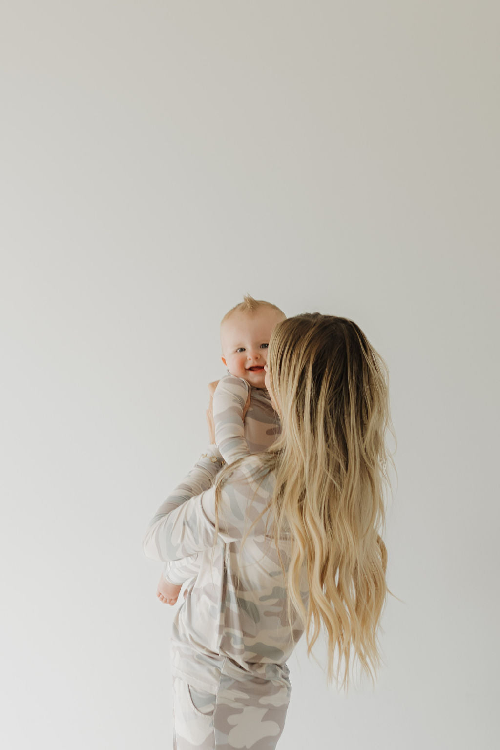 A woman with long blonde hair holds a smiling baby, both dressed in hypo-allergenic Bamboo Zip Pajamas in a light camo pattern by forever french baby. The plain white background accentuates their gentle matching attire, ideal for sensitive skin.