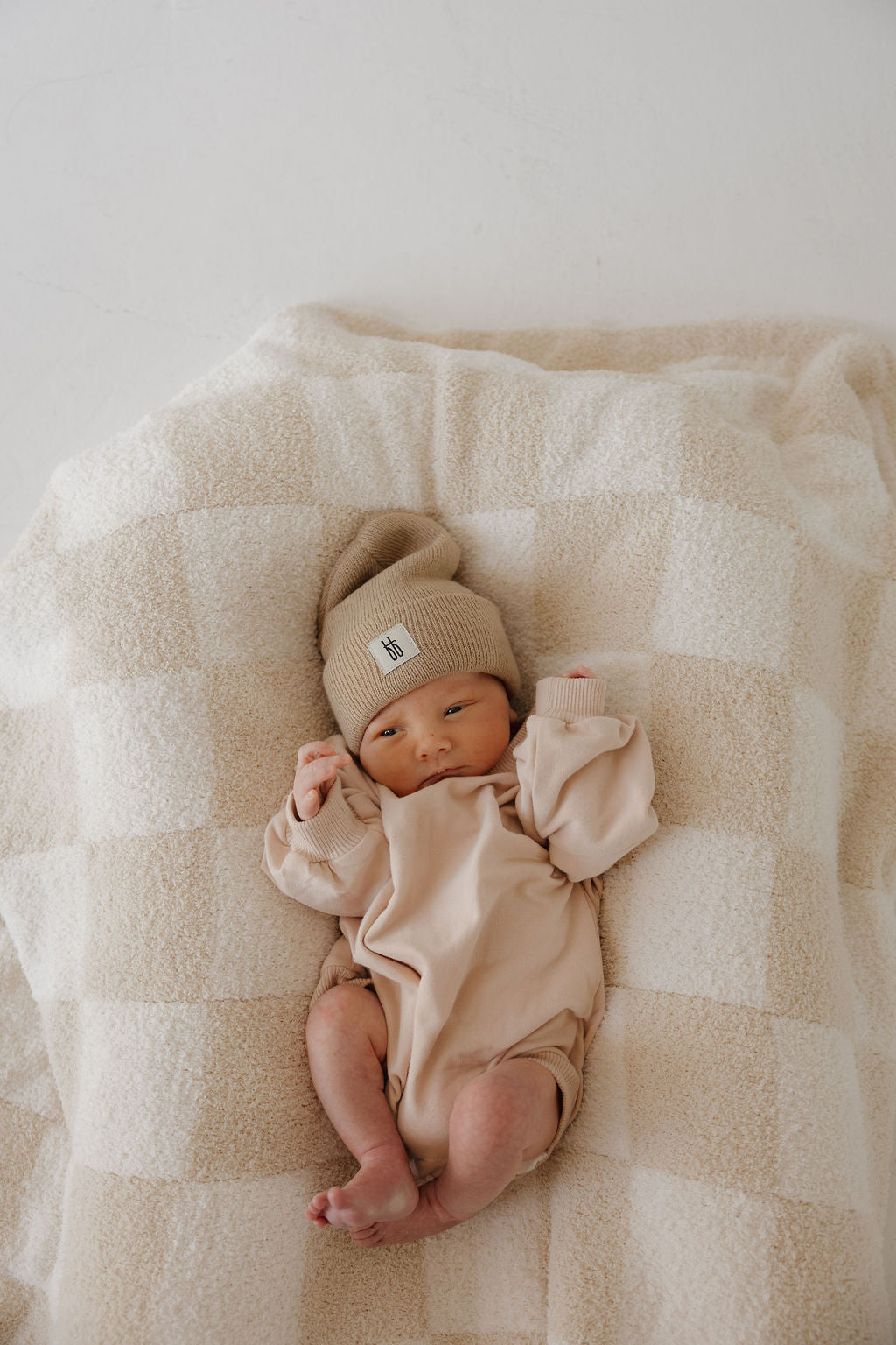 A newborn wearing the forever french baby Sweatshirt Romper in Cashew and a knit hat lies on a soft cream-and-white checkered blanket, looking up with hands near the face.