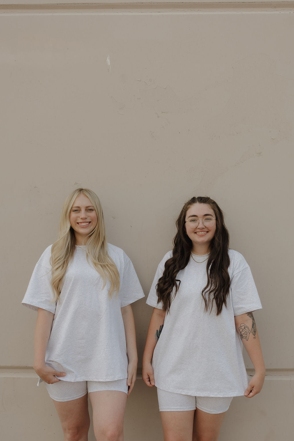 Two women smile side by side in forever french baby Women's Short Set | Heather—oversized tees and comfy shorts. One has long blonde hair; the other, long dark hair with glasses. Both look relaxed and happy against a beige wall.