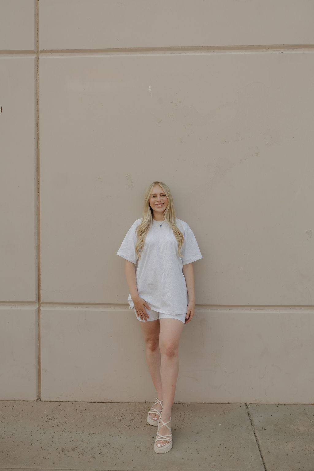 A woman with long blonde hair, wearing the Women's Short Set in Heather by forever french baby, stands smiling against a plain beige wall on a concrete sidewalk.