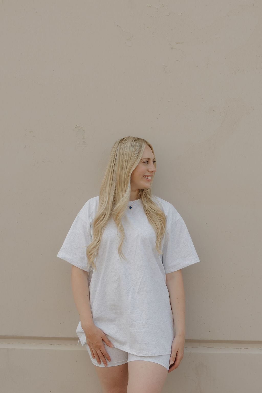 A woman with long blonde hair, wearing the forever french baby Women's Short Set in Heather, stands against a beige wall, smiling and looking to her left.
