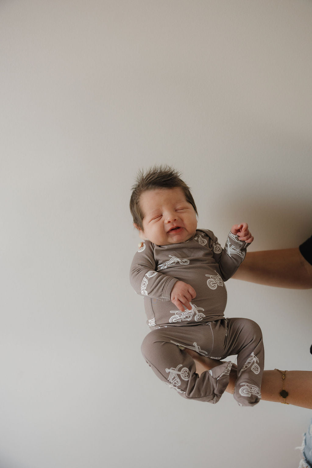 A baby in the forever french baby Sleepy Time Set | Dirt Bike, featuring brown bamboo fabric with white bike prints, is held sideways by an adult against a plain light wall, eyes closed and looking content.
