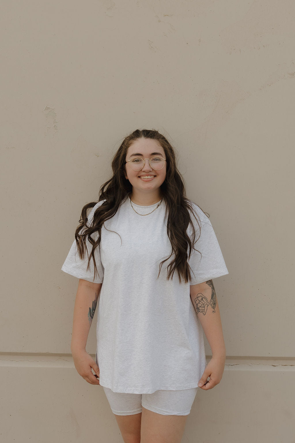 A woman with long brown hair and glasses smiles against a beige wall, wearing the forever french baby Women's Short Set | Heather. Her look is completed with visible forearm tattoos and layered necklaces.