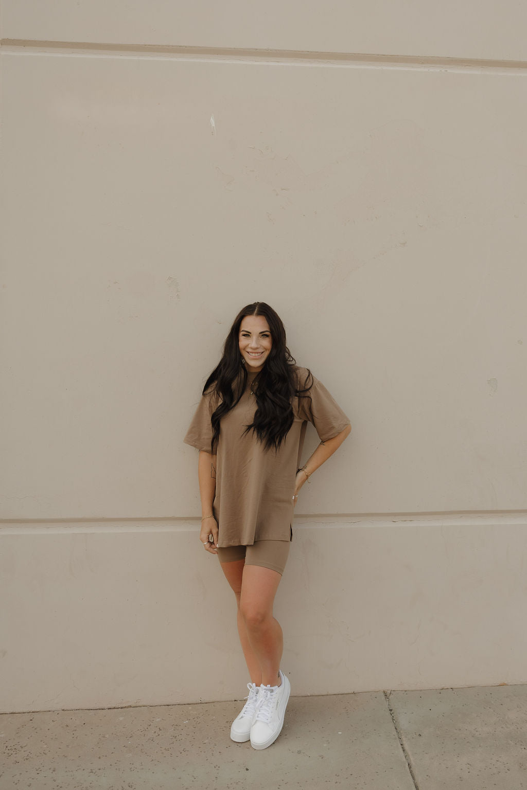 A woman with long dark hair stands confidently on a concrete surface, smiling in the forever french baby Women's Short Set | Patina and white sneakers, one hand on her hip, against a beige wall.