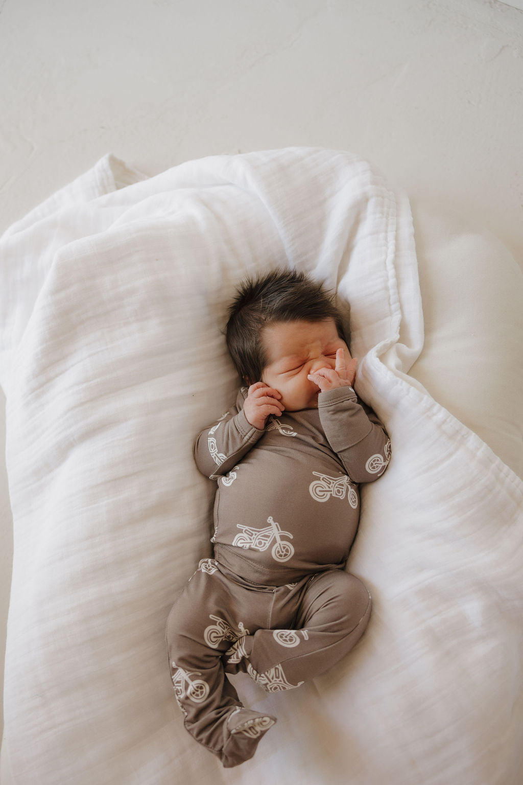 A newborn wearing the forever french baby Sleepy Time Set | Dirt Bike—a hypo-allergenic brown onesie with white motorcycle prints—lies peacefully on a cozy white blanket, eyes closed and hands by their face.