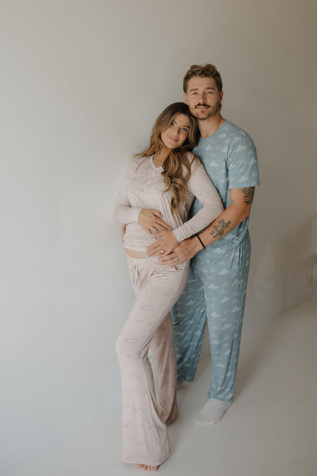 A couple stands together against a plain wall, both in Kendy 3 matching bamboo pajama sets. The woman cradles her baby bump while the man, wearing the Men's Short Sleeve Bamboo Pajama | Duke, embraces her and smiles in the soft, breathable fabric.