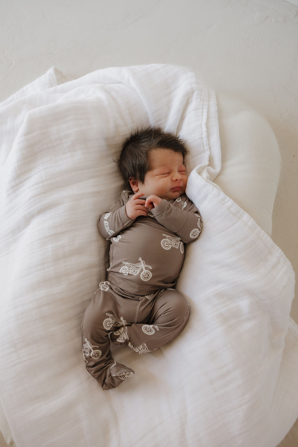 A newborn with dark hair sleeps on a soft white blanket, wearing the forever french baby Sleepy Time Set | Dirt Bike— a hypo-allergenic brown onesie with white motorcycle prints.