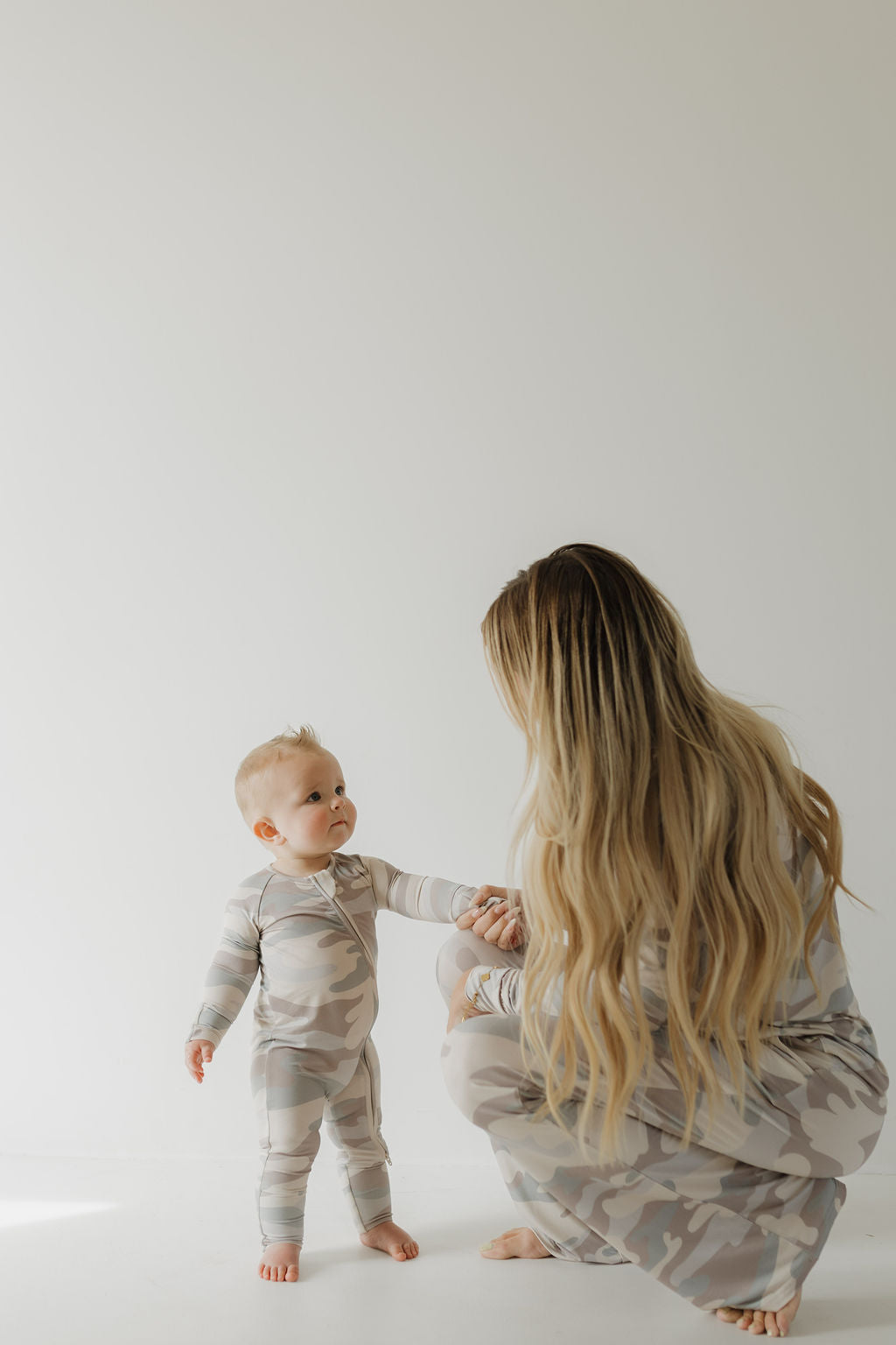 A mother and baby, both in matching "Women's Bamboo Pajama | Camo" outfits from forever french baby, are in a minimalist room. The mother crouches, gently holding the baby's hand as they stand together. Their hypo-allergenic clothing ensures comfort against the plain light-colored background.
