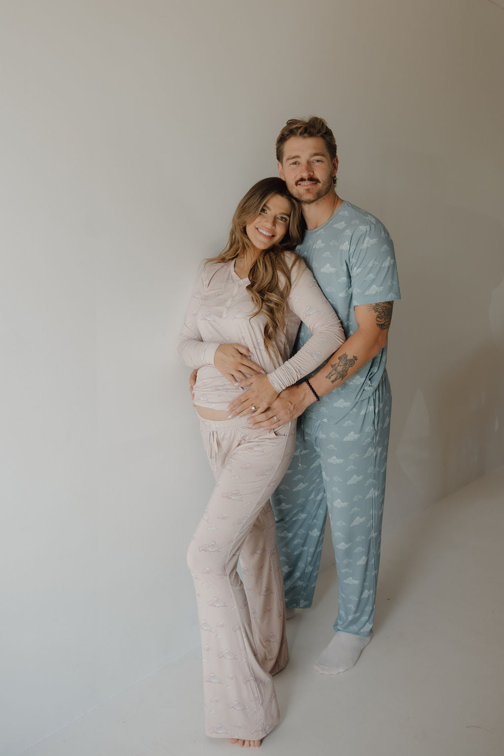 A pregnant woman in Kendy 3's Ruby Women's Bamboo Pajama stands smiling with a man in blue pajamas, his arm around her waist and her hand on her baby bump—ideal comfort for breastfeeding moms, set against a neutral background.
