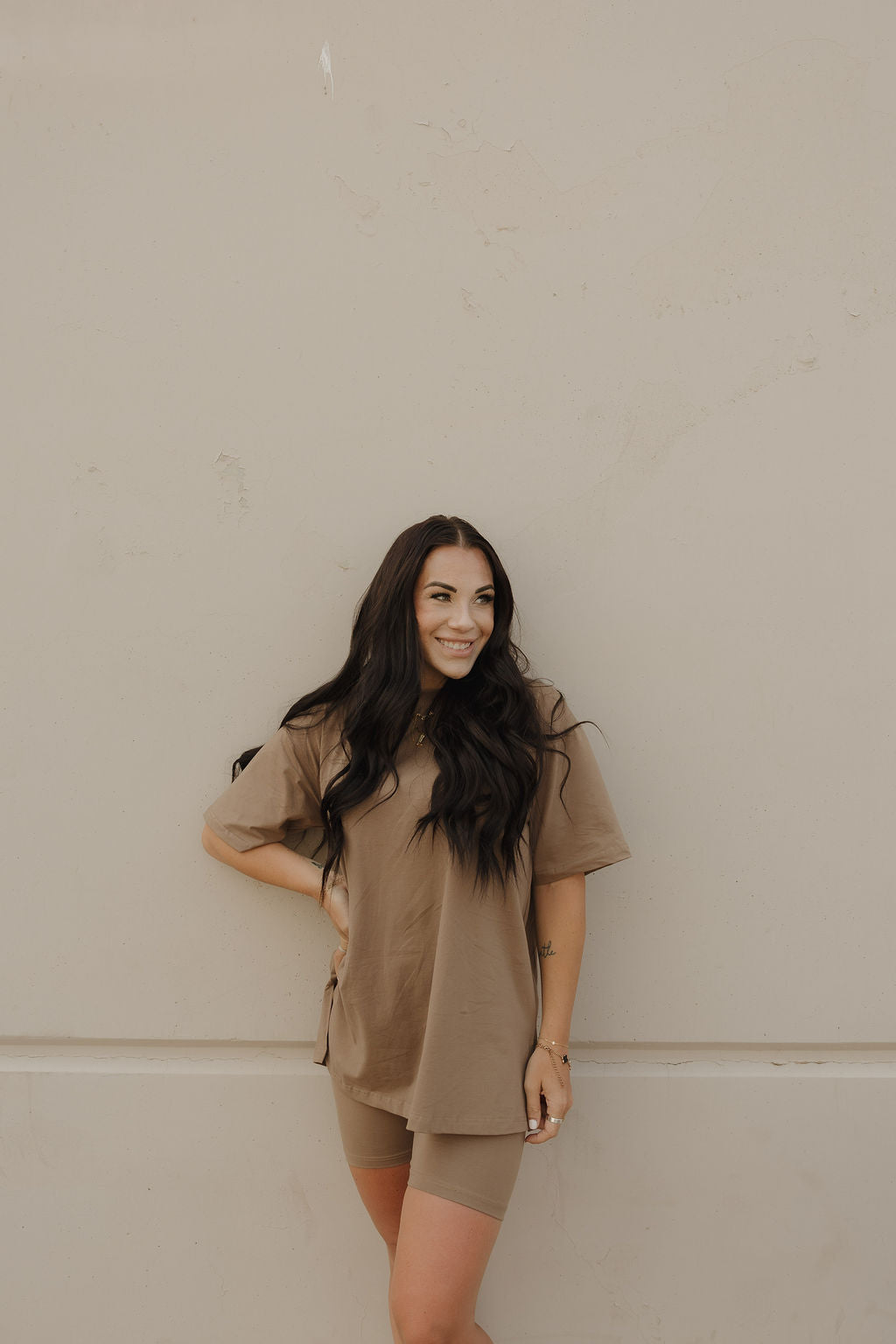 A woman with long dark hair smiles against a beige wall, wearing the forever french baby Women's Short Set in Patina, featuring an oversized tee and matching brown biker shorts with one hand on her hip.