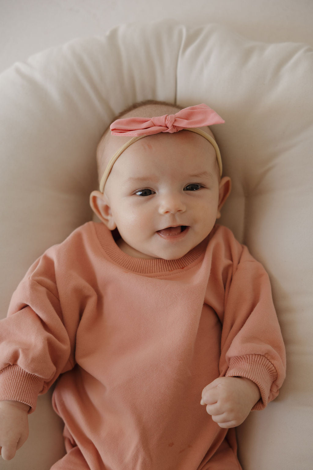 A baby in the forever french baby Sweatshirt Romper | Rose, wearing a pink bow headband, lies on a soft cream cushion and smiles gently at the camera.