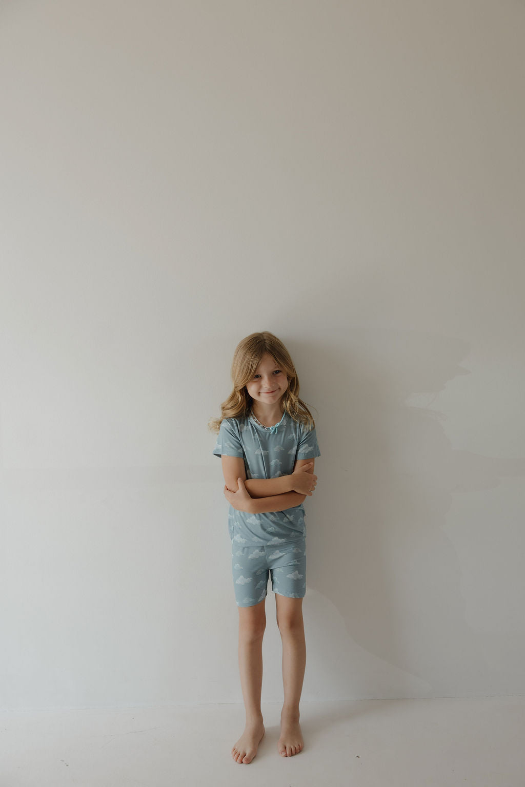 A young child with long blond hair stands barefoot against a light wall, wearing the Kendy 3 Bamboo Short Set | Duke—a soft, hypoallergenic, matching blue tee and shorts with a cloud pattern. The child has arms crossed and a neutral expression.