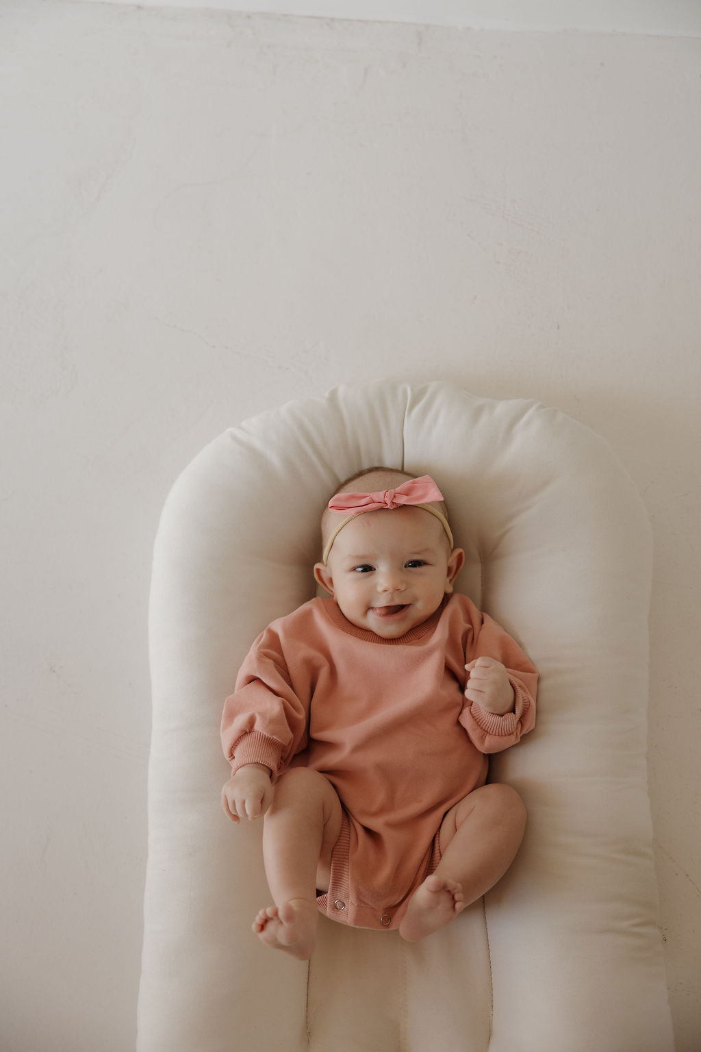 A smiling baby wearing the forever french baby Sweatshirt Romper | Rose lies on a soft white cushion, gazing up at the camera.