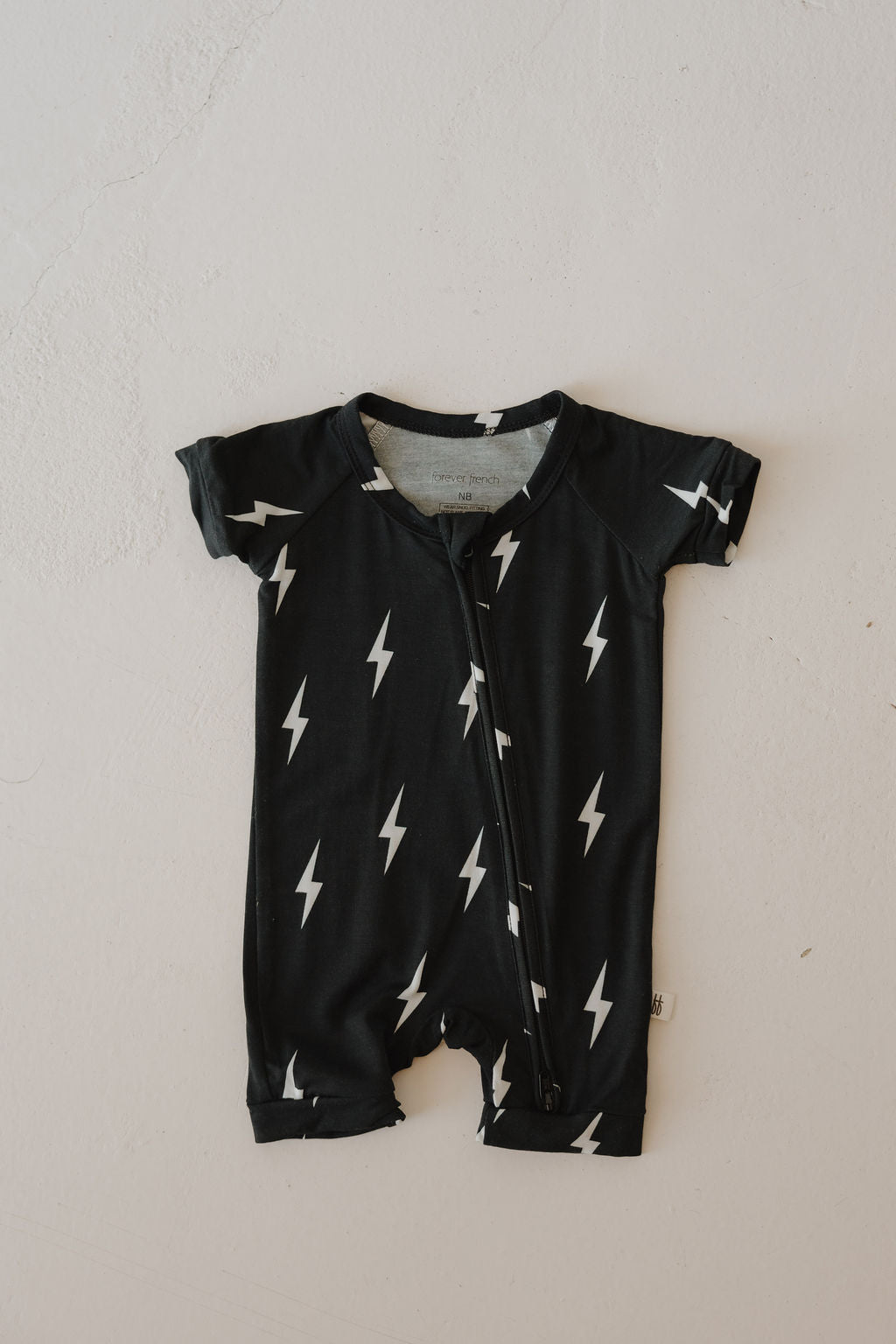 The Bamboo Zip Shortie in Midnight Lightning Bolt by forever french baby is a black short-sleeve romper with a diagonal zipper and white lightning bolt pattern, made from breathable bamboo fabric and shown laid flat on a light background.