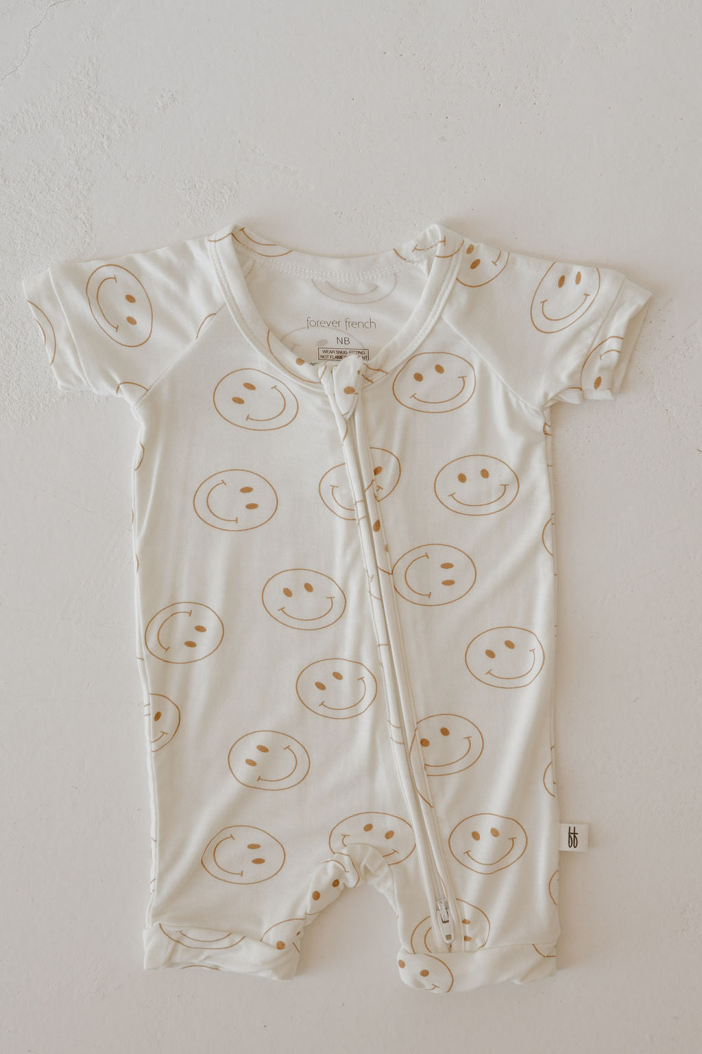 The forever french baby Bamboo Zip Shortie | Just Smile is a cream, short-sleeved romper with brown smiley faces and a front zipper, made from breathable fabric for all-day comfort.