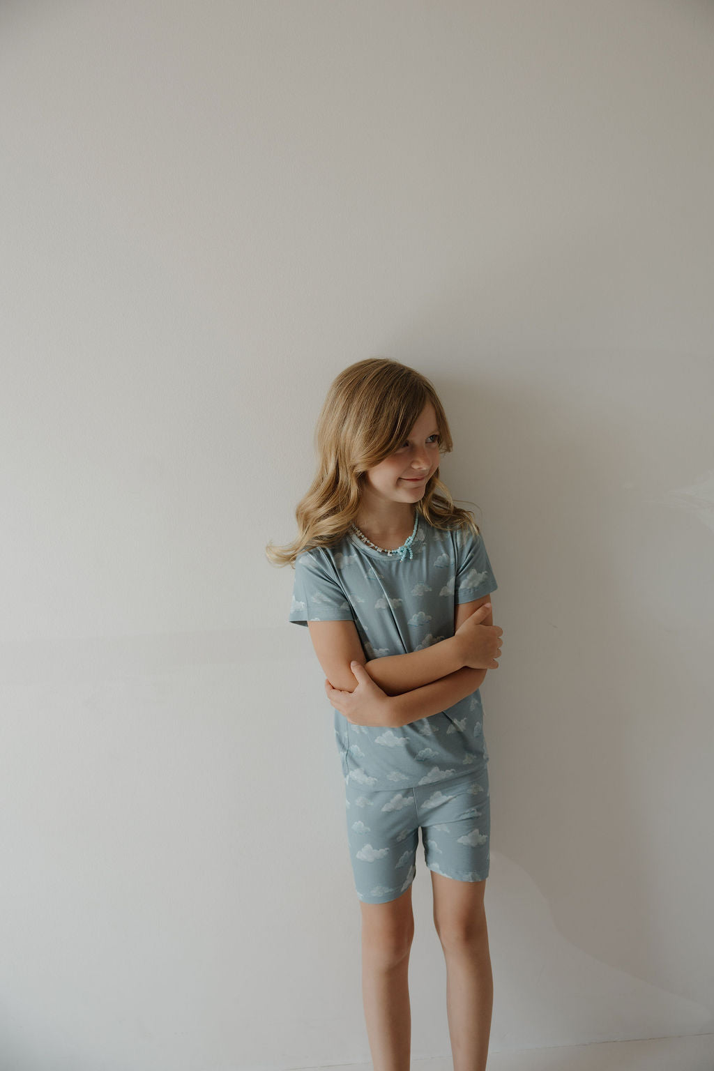 A young girl with long blonde hair stands against a plain light wall, smiling slightly with her arms crossed. She wears the Kendy 3 Bamboo Short Set | Duke—a hypo-allergenic, matching light blue T-shirt and shorts with a white cloud pattern.