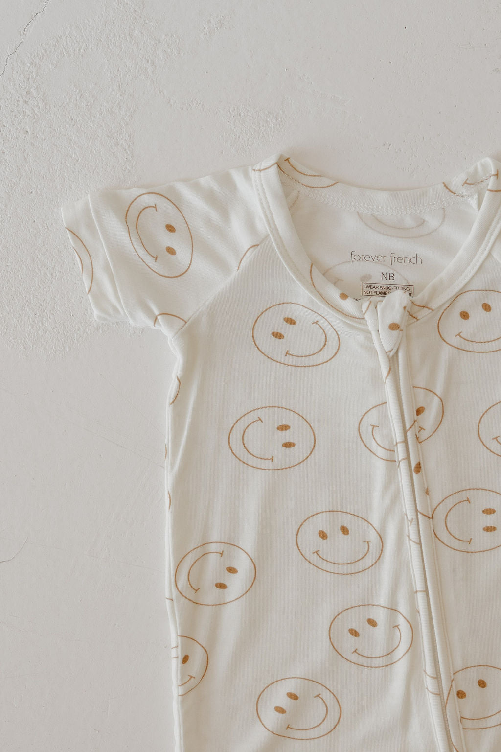 The Bamboo Zip Shortie | Just Smile by forever french baby is a cream short-sleeve onesie with light brown smiley faces, a front zipper, and "forever french" branding. Crafted from breathable fabric for baby comfort.