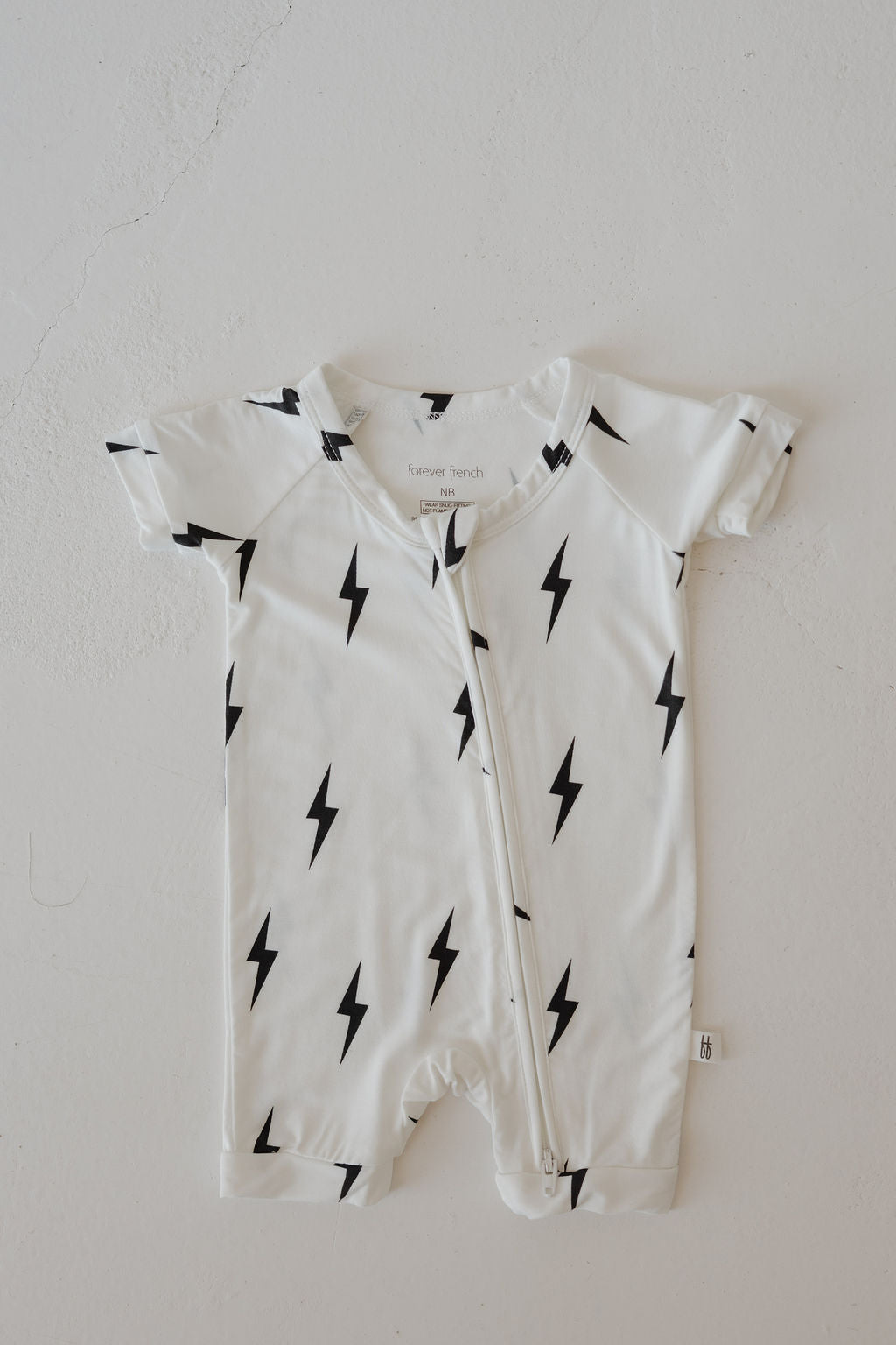 The forever french baby Bamboo Zip Shortie in White & Black Lightning Bolt, a hypo-allergenic bamboo romper with short sleeves and a diagonal zipper, is shown lying flat on a light surface.